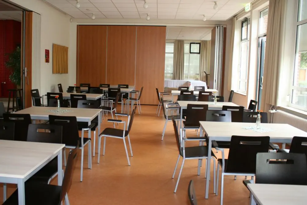 Foto van de zaal van de locatie 'De Kemphaan' Rechts ramen. Links en achterin een schuifwand. Links vier witte tafels met zwart/witte stoelen. Rechts drie tafels met dito stoelen.