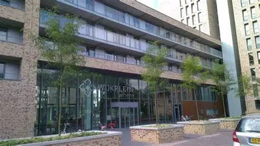 Foto van modern gebouw 'Wijkplein Where'. Op de voorgrond grote plantenbakken met jonge boompjes (en een stukje auto)