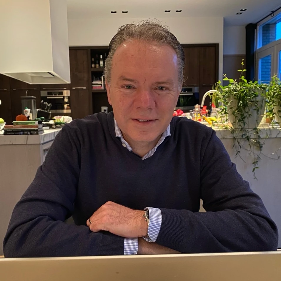 Mark Buitenhek. Hij draagt een donkere trui met daaronder een fijngeblokt overhemd. Op de achtergrond een keuken.